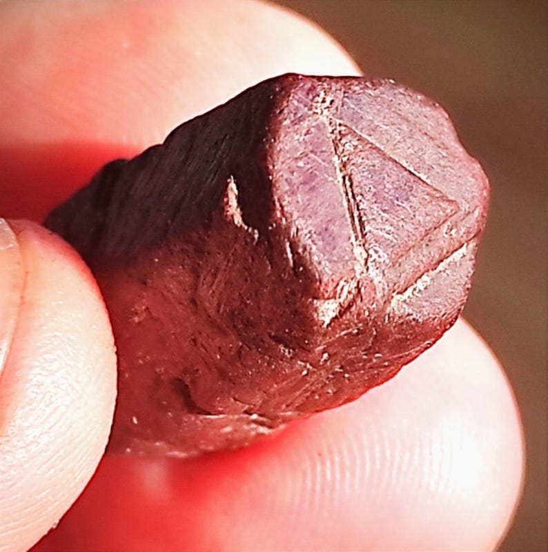 Op de afbeelding: Close-up van een ruwe, donkerrode edelsteen. De steen heeft een gefacetteerd oppervlak met een opvallende driehoekige vorm die in &eacute;&eacute;n kant is gesneden. De edelsteen wordt tussen vingers gehouden, wat de natuurlijke textuur en kleur benadrukt.