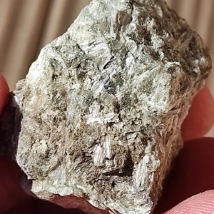Puede incluir: Primer plano de una roca rugosa, gris y verde con textura cristalina. La roca tiene un brillo plateado y parece estar compuesta por muchos cristales pequeños y planos.