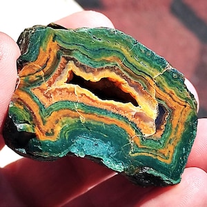 Puede incluir: Una sección transversal de una geoda, que muestra capas de tonos verdes, naranjas y amarillos. La piedra tiene un centro hueco, revelando una cavidad. La formación rocosa natural muestra un patrón de bandas, con un exterior rugoso y texturizado.