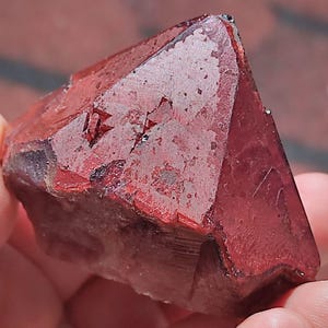 May include: A deep red, faceted mineral specimen with a triangular, pyramid-like shape. The surface displays a textured, crystalline structure with sharp edges and natural indentations, showcasing a rich, opaque crimson hue throughout the stone.
