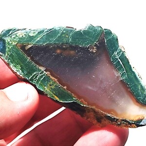 Può includere: Una fetta di pietra preziosa lucidata con un bordo esterno verde scuro e un centro traslucido di colore chiaro. La pietra ha una consistenza ruvida e naturale ed è tenuta in mano, mostrando i suoi motivi e colori unici.