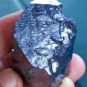 Könnte beinhalten: Dunkles, metallisches Mineralexemplar mit facettierter, kristalliner Struktur. Die Oberfläche reflektiert Licht und zeigt eine Reihe von Blau- und Silbertönen. Die grob behauenen Kanten und die einzigartige Form unterstreichen seine natürliche Form.