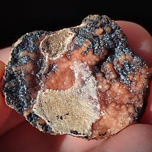 Op de afbeelding: Close-up van een natuurlijk mineraal specimen. De steen heeft een mix van texturen en kleuren, waaronder gebieden in donkergrijs, perzik en bruin. Het oppervlak heeft op sommige plaatsen een bubbelachtig, kristallijn uiterlijk.