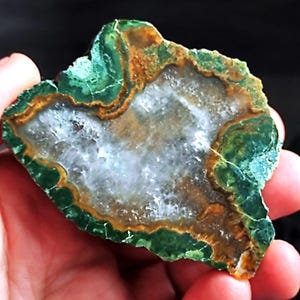 Può includere: Un campione di pietra naturale grezza con un centro di geode. La pietra presenta uno strato esterno verde vibrante con bande marroni e dorate, che circondano un interno di cristallo bianco scintillante. La pietra è tenuta in una mano.