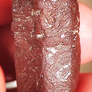 Op de afbeelding: Een close-up van een donkerrode, rechthoekige edelsteen met een ruw, getextureerd oppervlak. De edelsteen heeft een enigszins onregelmatige vorm en lijkt een ruwe, ongeslepen steen te zijn. De kleur is een diep, rijk rood, met lichtere plekken.