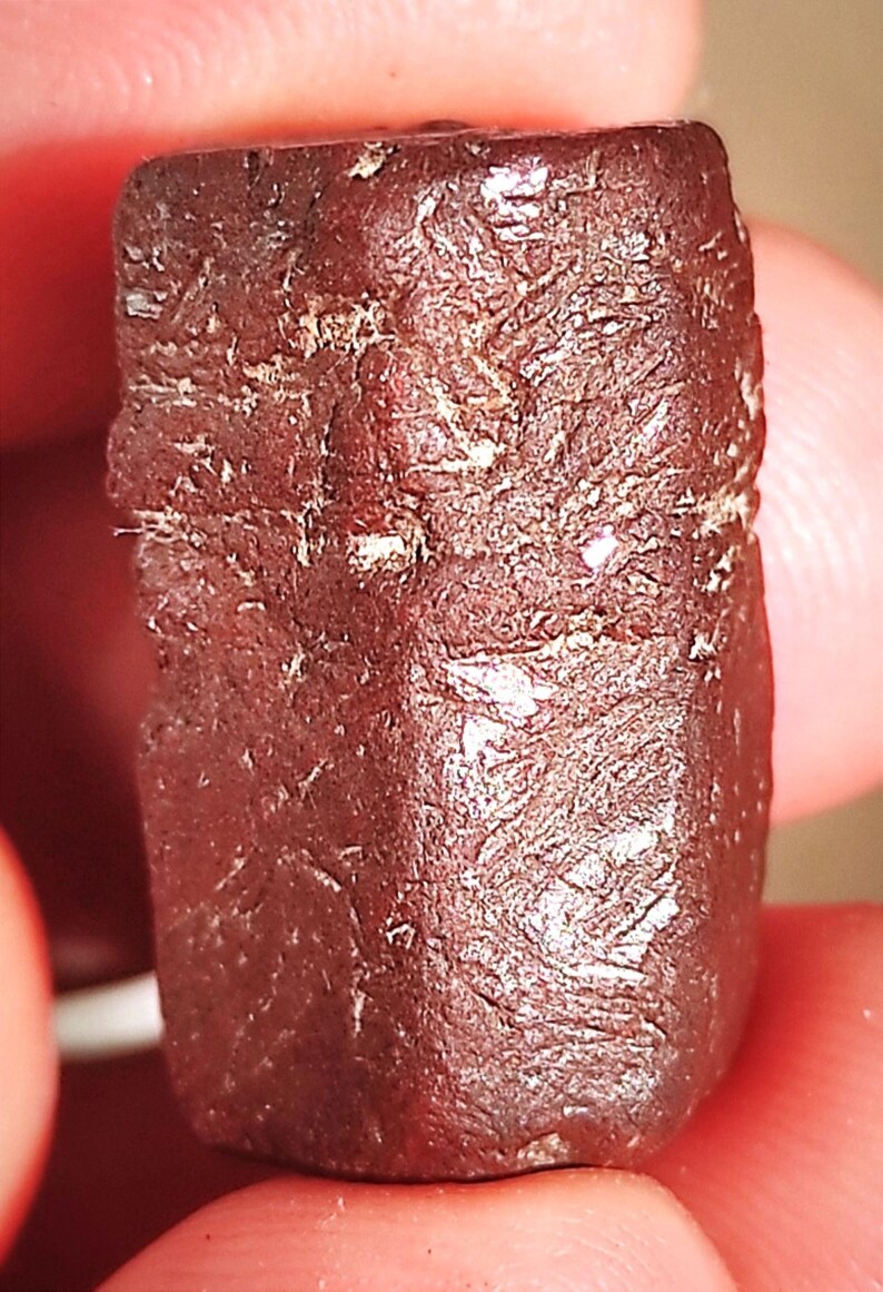 Op de afbeelding: Een close-up van een donkerrode, rechthoekige edelsteen met een ruw, getextureerd oppervlak. De edelsteen heeft een enigszins onregelmatige vorm en lijkt een ruwe, ongeslepen steen te zijn. De kleur is een diep, rijk rood, met lichtere plekken.