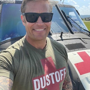 May include: A man wearing sunglasses and a green t-shirt with a red cross and the text "DUSTOFF" stands in front of a black helicopter.