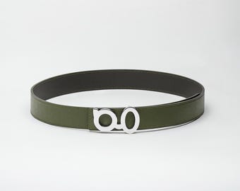 May include: An olive green belt with a silver-colored buckle. The belt is made of a smooth material and has a dark brown interior. The buckle features a stylized design.