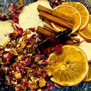 Puede incluir: Arreglo aromático con rodajas de naranja secas, rodajas de manzana, palitos de canela y una pequeña botella de vidrio marrón con cuentagotas. La composición incluye pétalos de rosa secos y otros elementos botánicos.