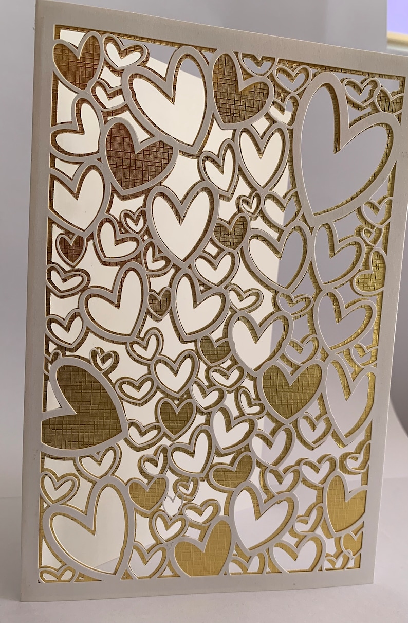May include: A white card with a gold background and a cut-out design of many hearts. The hearts are layered and overlap, creating a complex and intricate pattern.