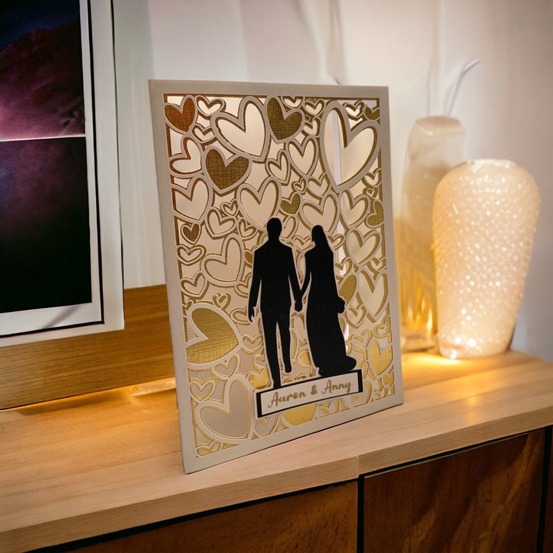 May include: A white card with a gold and white heart design. The card features a silhouette of a couple holding hands. The text "Aaron & Amy" is printed on the card.