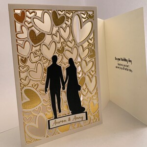 May include: A white card with a gold heart pattern and a silhouette of a couple holding hands. The card is open to reveal a white interior with the text "On your Wedding day may love be in your heart, and every joy fill this day."