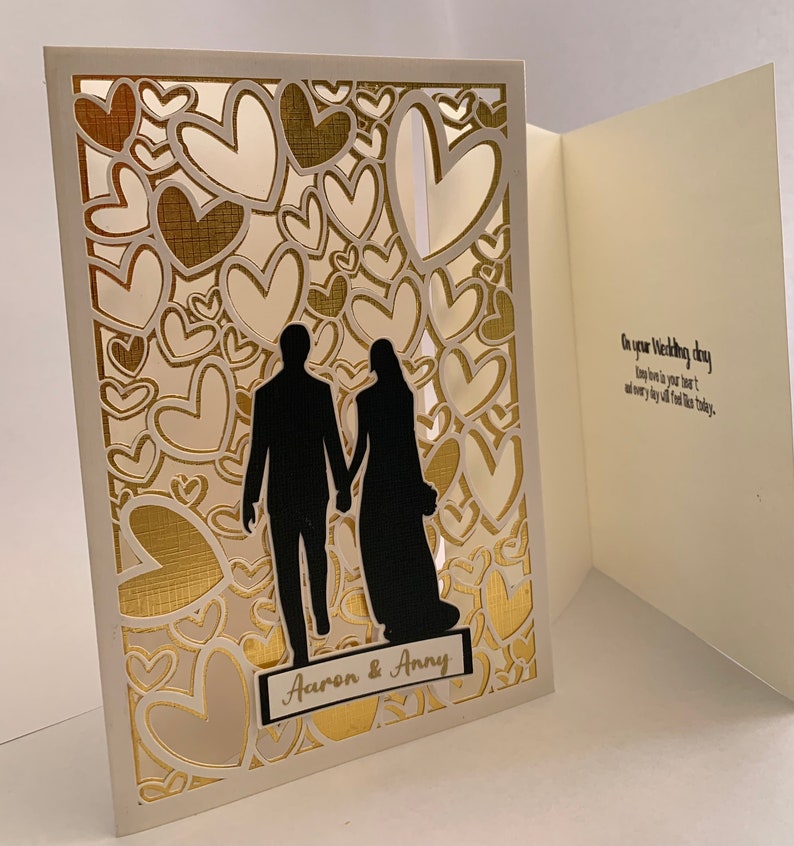 May include: A white card with a gold heart pattern and a silhouette of a couple holding hands. The card is open to reveal a white interior with the text "On your Wedding day may love be in your heart, and every joy fill this day."