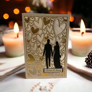 May include: A white card with a gold and white heart pattern. The card features a silhouette of a couple standing together. The text "Aaron & Amy" is printed on the card.