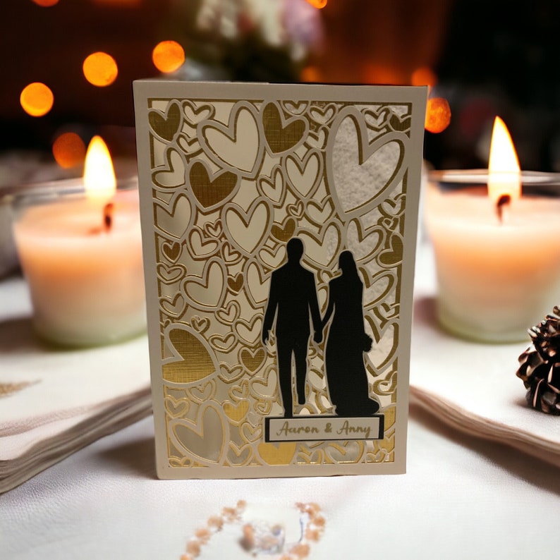 May include: A white card with a gold and white heart pattern. The card features a silhouette of a couple standing together. The text "Aaron & Amy" is printed on the card.