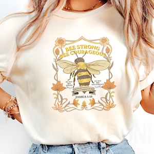 Puede incluir: Camiseta color crema con una ilustración detallada de una abeja y el texto "BEE STRONG BEE COURAGEOUS" en un marco decorativo. El diseño incluye elementos florales y la referencia bíblica Josué 1:19.