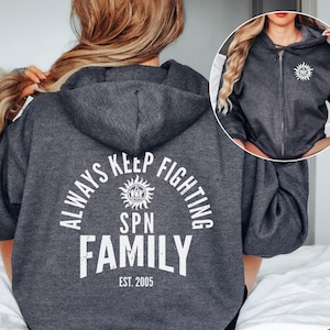 Może przedstawiać: Ciemnoszara bluza z kapturem i zamkiem błyskawicznym z kapturem i białym napisem na plecach: "ALWAYS KEEP FIGHTING SPN FAMILY EST. 2005". Mniejszy obrazek przedstawia przód bluzy z grafiką słońca.