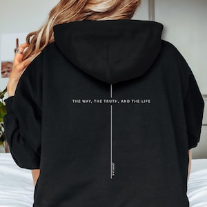Könnte beinhalten: Schwarzer Kapuzenpullover mit weißem Text auf der Rückseite. Der Text lautet "THE WAY, THE TRUTH, AND THE LIFE" mit einer vertikalen Linie und "JOHN 14:6" darunter. Die untere rechte Ecke des Hoodies hat einen schwarzen Kreis mit dem Wort "BLACK" in Weiß.