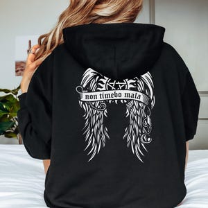 May include: Black hoodie with a white graphic of angel wings and the text "non timebo mala". The hoodie has a drawstring hood and a kangaroo pocket. The word "BLACK" is in a white circle at the bottom right.