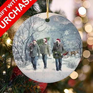 May include: Round Christmas ornament featuring three people walking in a snowy forest, wearing Santa hats. The ornament has the words "Merry Christmas" and "Happy New Year!" on it. A red banner reads "GIFT WITH PURCHASE".