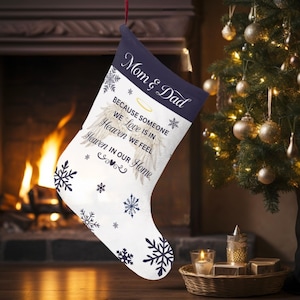 May include: White Christmas stocking with a navy blue cuff, decorated with snowflakes and the text "Mom & Dad". The stocking includes the message "Because someone we love is in Heaven we feel Heaven in our Home".