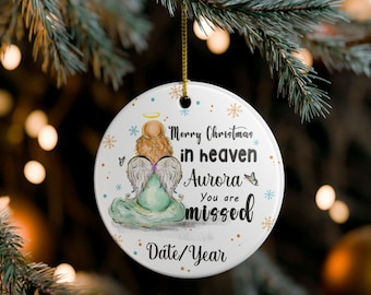 Op de afbeelding: Ronde kerstversiering met een aquarel illustratie van een engel in een groene jurk, met de tekst "Merry Christmas in Heaven Aurora You are missed Date/Year". De witte versiering heeft gouden accenten en een gouden draad.