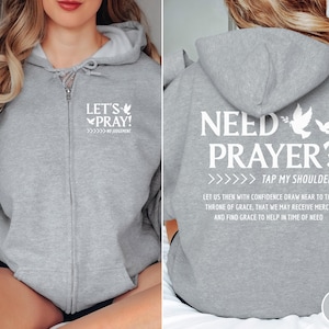 Könnte beinhalten: Grauer Kapuzenpullover mit Reißverschluss. Auf der Vorderseite steht in Weiß "LET'S PRAY!", auf der Rückseite "NEED PRAYER? TAP MY SHOULDER". Der Hoodie hat eine Kapuze und einen durchgehenden Reißverschluss.