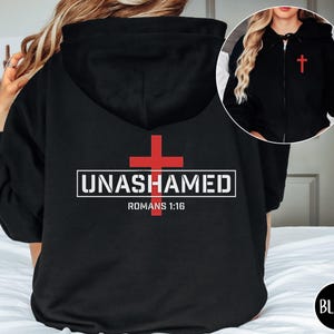 Könnte beinhalten: Schwarzer Kapuzenpullover mit Reißverschluss, einem roten Kreuz und dem Text "UNASHAMED" und "ROMANS 1:16" auf der Rückseite.