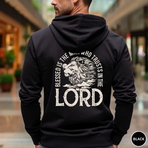Könnte beinhalten: Schwarzer Kapuzenpullover mit weißem Aufdruck auf dem Rücken. Der Aufdruck zeigt einen Löwenkopf und die Worte "Blessed is the man who trusts in the Lord". Das Wort "LORD" ist in großen, fetten Buchstaben geschrieben. Unten rechts befindet sich ein schwarzer Kreis mit dem Wort "BLACK".