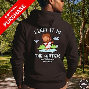 Könnte beinhalten: Dunkelbrauner Kapuzenpullover mit dem Text "I LEFT IT IN THE WATER" und einer Illustration von Jesus. Auf der Rückseite des Hoodies steht außerdem der Text "MATTHEW 28:19" und das Datum "09-07-2025".