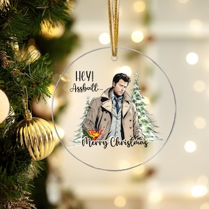 Könnte beinhalten: Transparenter, runder Weihnachtsschmuck mit goldenem Band. Der Schmuck zeigt eine Cartoon-Illustration eines Mannes in einem beigefarbenen Mantel mit dem Text "HEY! Assbutt... Merry Christmas". Zwei grüne Weihnachtsbäume sind hinter dem Mann.