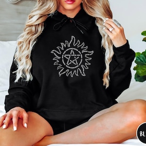 Może przedstawiać: Czarna bluza z kapturem z białym nadrukiem. Projekt przedstawia gwiazdę w okręgu, otoczoną tekstem. Bluza ma kaptur ze sznurkiem i jest wykonana z miękkiego materiału.