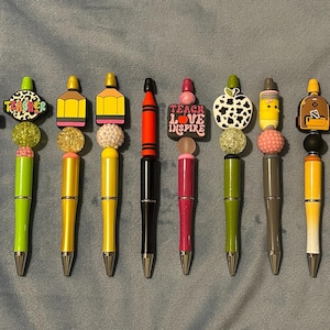 May include: A collection of colorful pens with decorative toppers. The toppers feature various school-themed designs, including a leopard print notebook, a crayon, an apple, and the words "TEACH LOVE INSPIRE". The pens have beaded accents and silver tips.