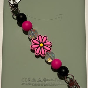 May include: A pink and black beaded phone strap with a flower charm. The strap has a heart-shaped clip on each end. The strap is attached to a green Kindle e-reader.
