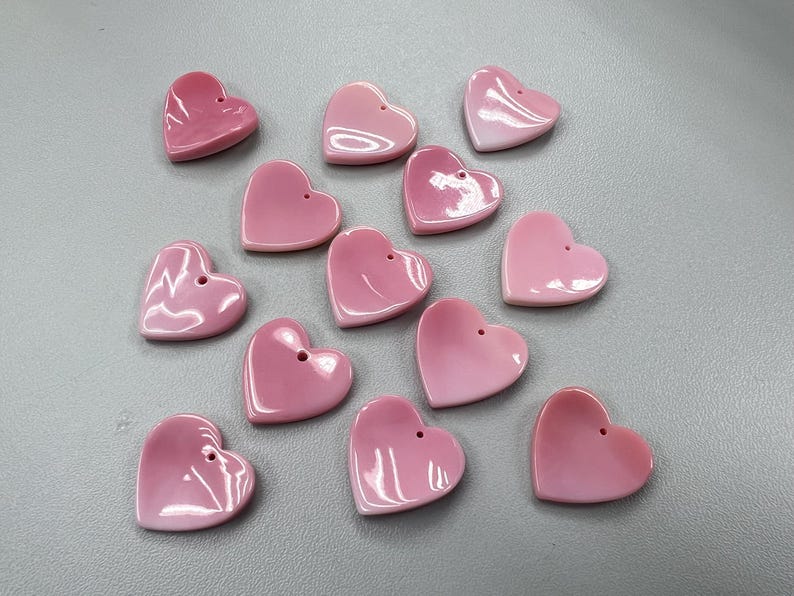 Puede incluir: Una colecci&oacute;n de 13 dijes de concha de coraz&oacute;n rosa con un agujero en la parte superior para colgar.