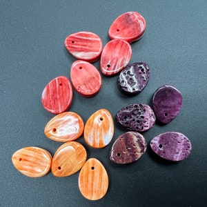 May include: Assortment of 12 colorful oval-shaped gemstone pendants with holes for stringing. The pendants are in shades of pink, orange, purple, and brown.