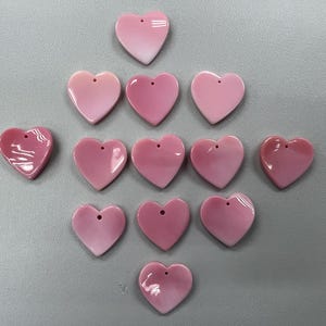 Puede incluir: Una colecci&oacute;n de 12 dijes de concha de coraz&oacute;n rosa con un agujero en el centro. Los dijes est&aacute;n dispuestos en un patr&oacute;n triangular sobre una superficie gris.