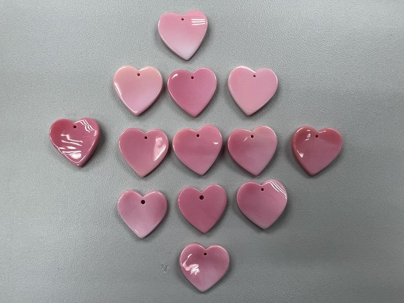 Puede incluir: Una colecci&oacute;n de 12 dijes de concha de coraz&oacute;n rosa con un agujero en el centro. Los dijes est&aacute;n dispuestos en un patr&oacute;n triangular sobre una superficie gris.
