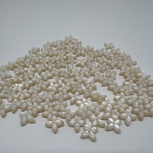 Puede incluir: Una gran cantidad de cuentas blancas en forma de flor, con aspecto nacarado. Las cuentas están esparcidas sobre una superficie blanca.