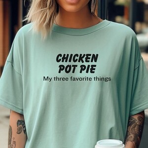 May include: A woman wearing a light blue t-shirt with the text "Chicken Pot Pie My three favorite things" in black. She is holding a to-go coffee cup.