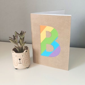 May include: A brown notebook with a rainbow letter "B" on the front. The notebook is sitting on a white surface next to a small, beige bear-shaped planter with a succulent plant.