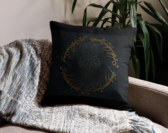 May include: Black square throw pillow featuring a gold ring inscription and a tree design. The pillow is on a gray sofa with a blue and white patterned blanket. Decorative home decor.