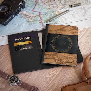Può includere: Porta passaporto in pelle nera con design a mappa e stemma dorato. Il porta passaporto è aperto, rivelando un passaporto e carte di credito. Sono visibili anche una fotocamera nera e un orologio con cinturino in pelle marrone.