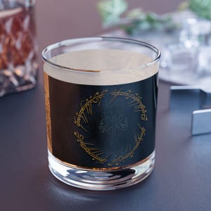 Puede incluir: Un vaso de vidrio transparente con un líquido oscuro y espuma. El vaso presenta una etiqueta negra con letras doradas y un diseño de árbol, que recuerda a un tema de fantasía. El vaso está sobre una superficie oscura.