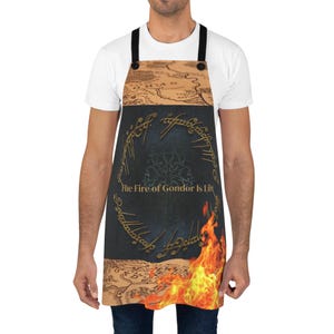 Op de afbeelding: Schort met een kaartontwerp en de tekst "The Fire of Gondor Is Lit". De schort heeft een donkerblauwe achtergrond met een boom- en vuurmotief. De schort heeft zwarte bandjes en knopen.