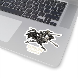 May include: A die-cut sticker featuring a black and white illustration of a skeleton wearing a hat and riding a large rooster while holding a long spear. The skeleton points forward with one hand. Below the illustration, the text ESPOLÓN TEQUILA is printed in a light beige color.