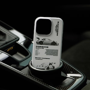 以下が含まれることがあります： 白いスマートフォンケース。黒いポルシェ911 GT3 RSのグラフィックが描かれ、「SANTORINI」、「PORSCHE 911 GT3 RS」、「GRAND TOURISMO」、「RENNSPORT」という文字が保護カバーの背面に黒色でプリントされています。