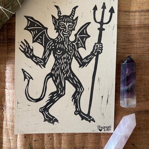 Peut inclure: Une impression rectangulaire sur papier beige clair présentant une illustration en linogravure à l'encre noire d'un diable cornu et ailé tenant un trident. Le texte "strangel knot" avec un petit cœur est imprimé dans le coin inférieur droit.