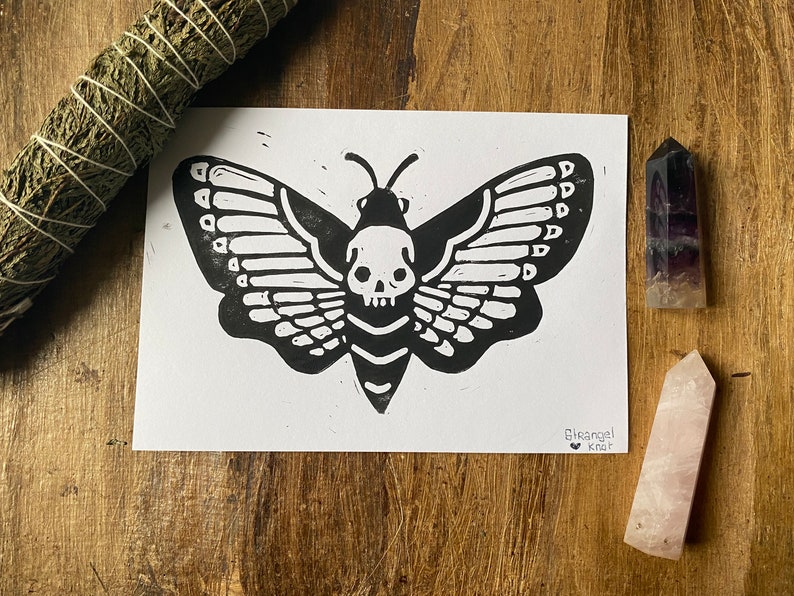 May include: Black and white print of a moth with a skull for a head. The print is on white paper and has the text "Blackangel Knit" in the bottom right corner.