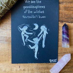 以下が含まれることがあります： 黒い紙に白黒のプリントが施され、「We are the granddaughters of the witches You couldn't burn」というテキストと、三日月を囲んで踊る3人の人物のスタイリッシュな画像が描かれています。プリントは右下に「Strangel Knot」と署名されています。